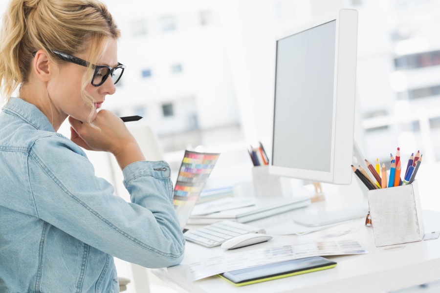 Woman wearing glasses sits at a desk looking at a computer screen while holding a pen, appearing focused as she reviews work related to managing or fixing issues like stolen pins on Pinterest.